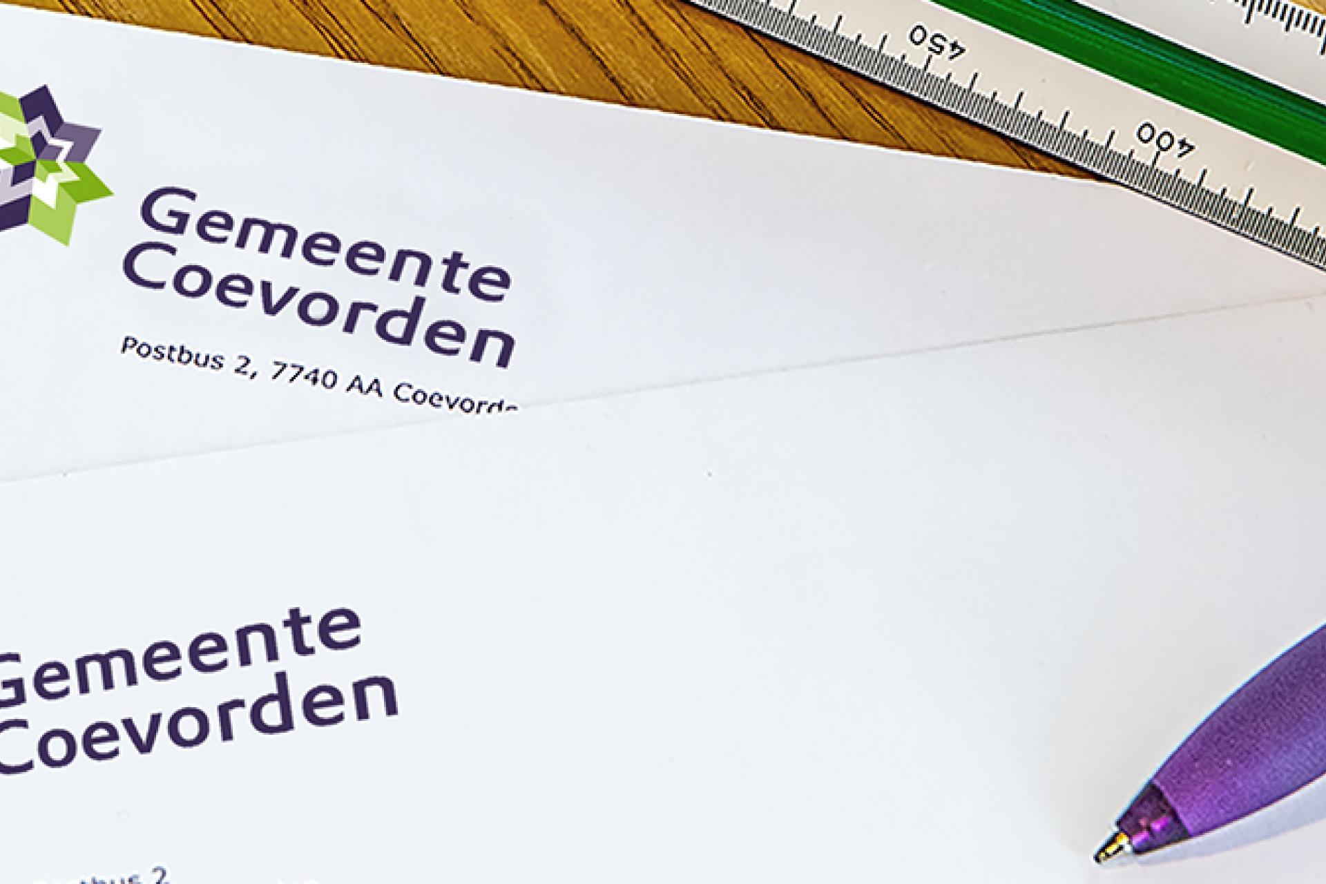 Stempels, enveloppen met logo gemeente Coevorden, een paarse balpen en meetlat vormen benodigdheden voor Belastingen op een houten achtergrond.