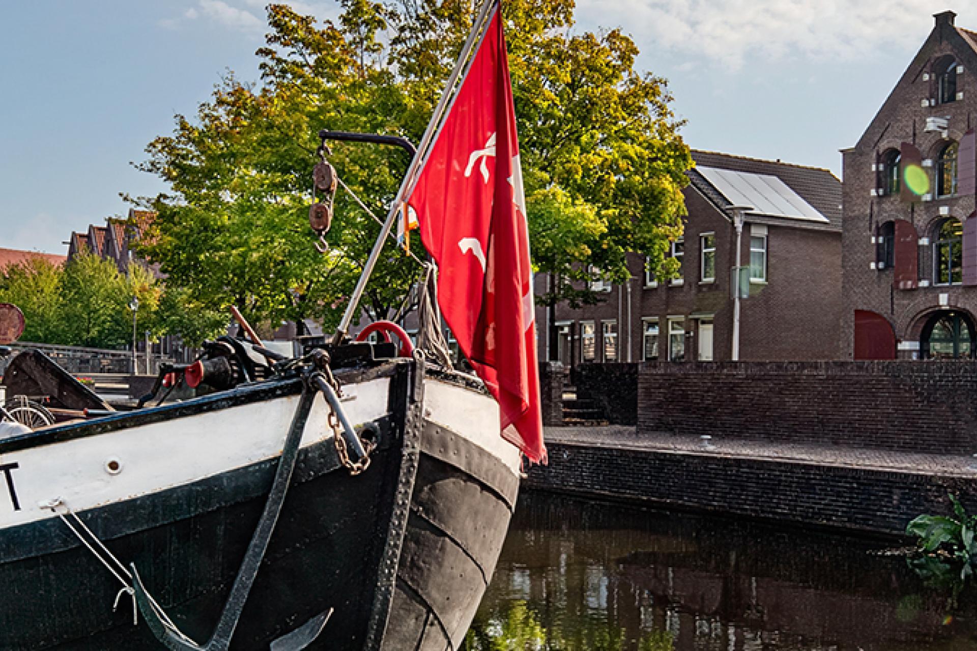 Op de voorgrond schip met rode vlag in haven, rechts 3 pakhuizen, nu stedelijk museum. Links voetpad langs het water. Op de achtergrond groene bomen en blauwe lucht.