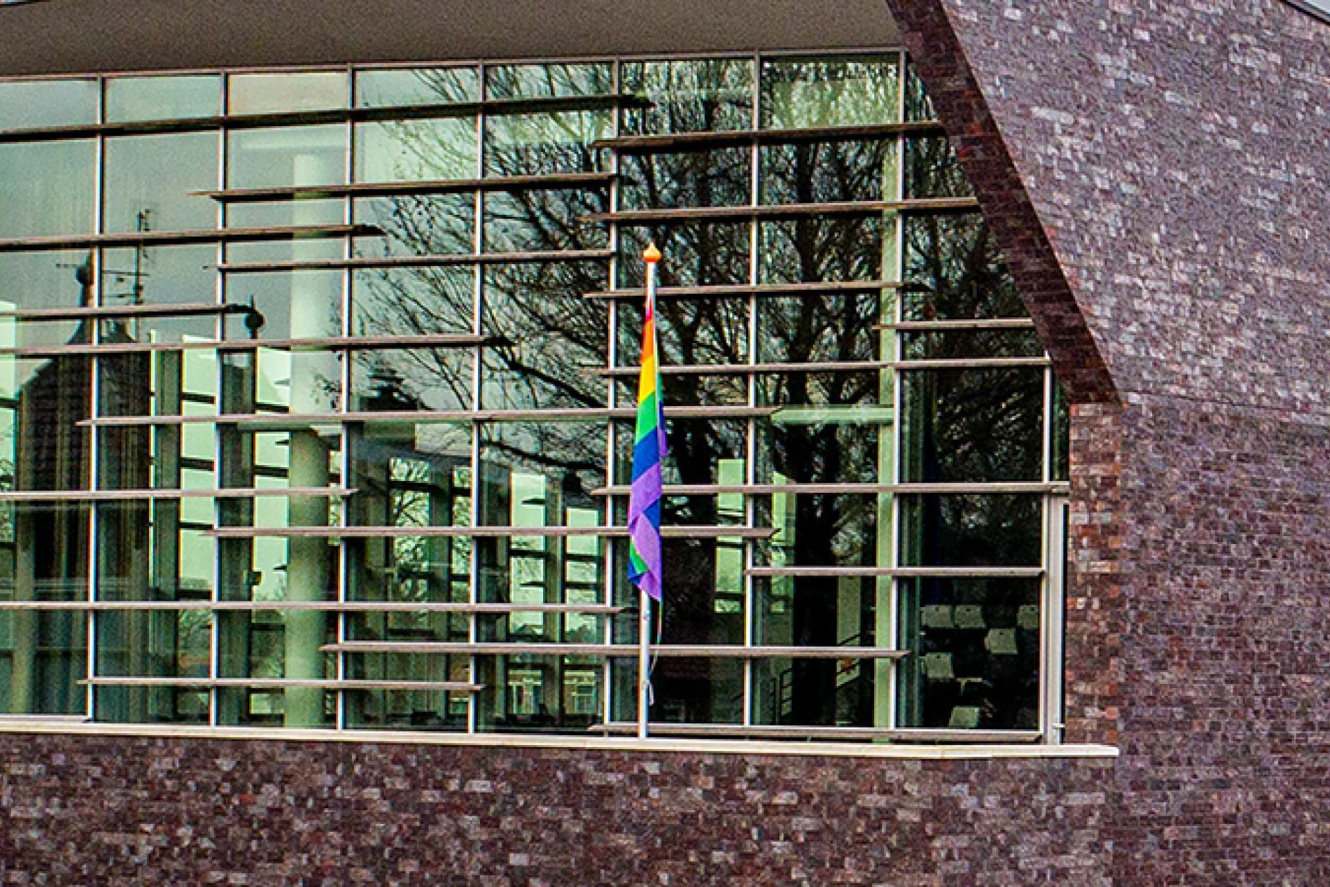 Zijaanzicht glazen pui van het gemeentehuis met aan de rechterkant rij vlaggen en een kale boom 